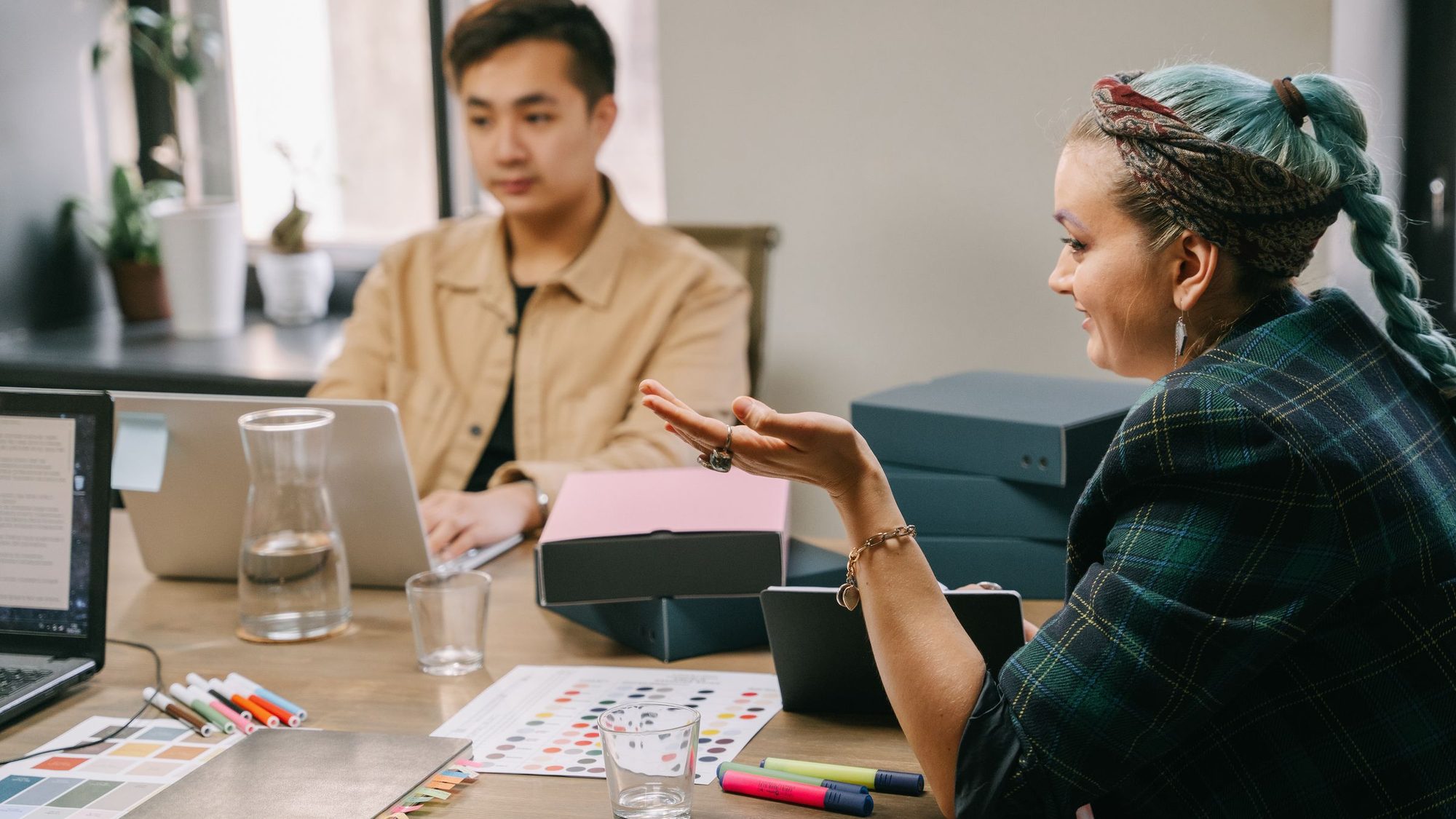 Two coworkers talk at a desk covered with color swatches and markers