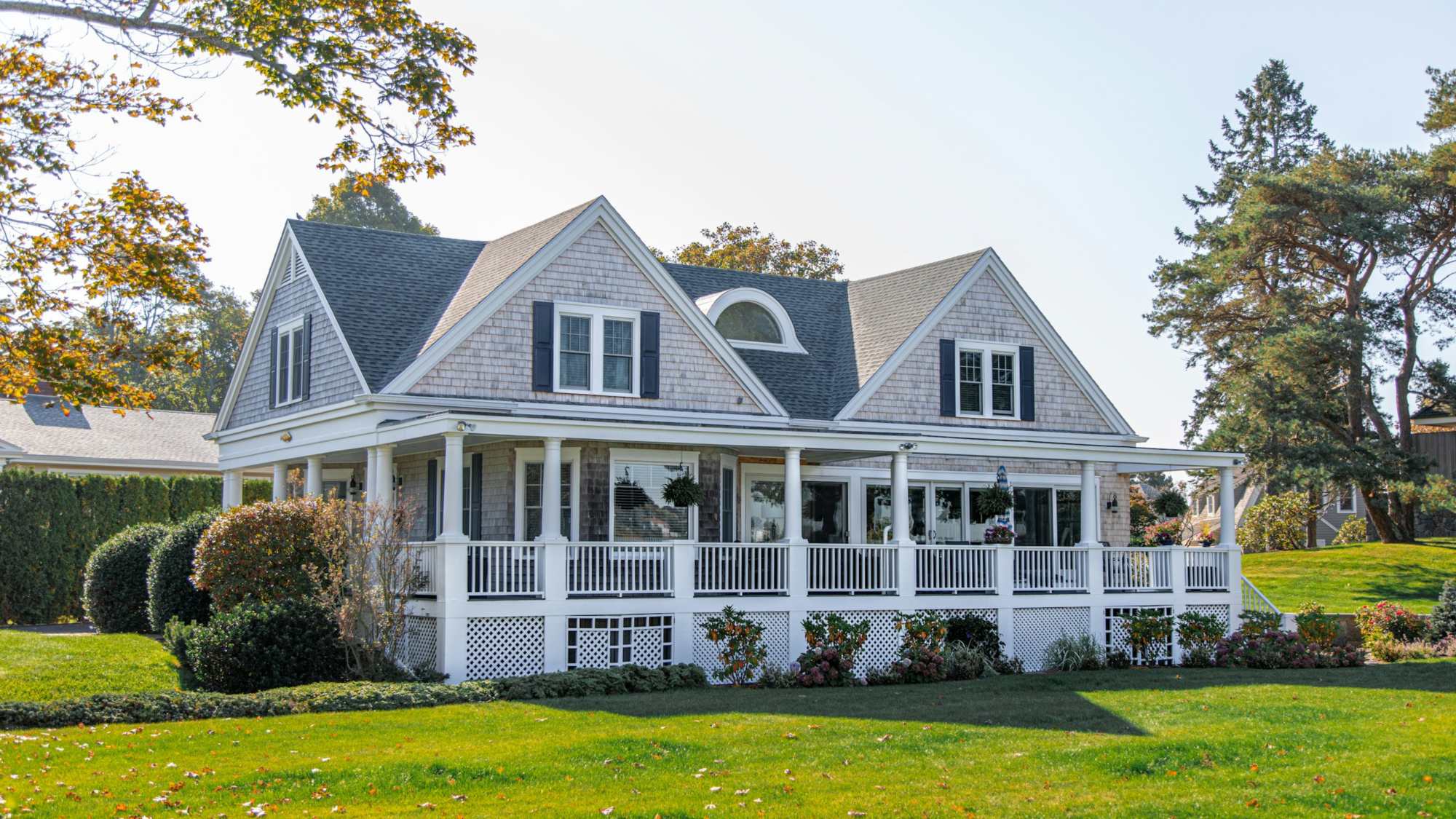A large gray shingled house with a white porch and a green lawn