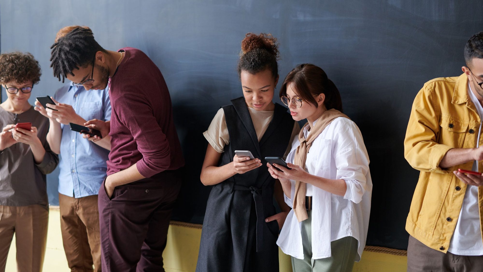 A group of young adults stand against a wall, each looking at a phone