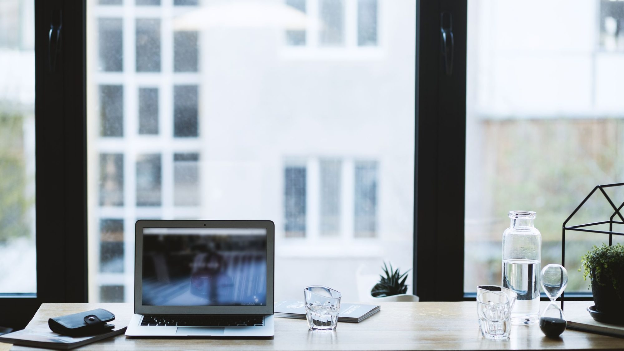 A laptop on a desk by a large window with a glass of water and a plant
